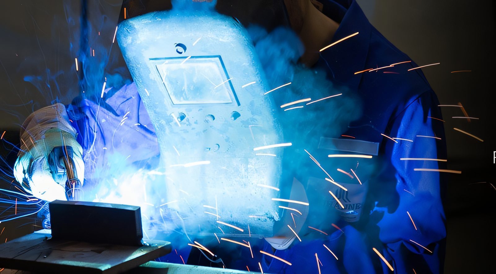 A welder in a blue jacket behind the mask, the arc lighting up the smoke and throwing sparks over the workpiece